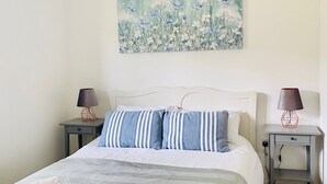 A neatly made bed with a striped pillow and a folded towel, a painting of flowers on the wall, and two bedside tables with lamps. - Beautiful, stylish New Forest 2 bed cottage, close to the beach! (Lymington)