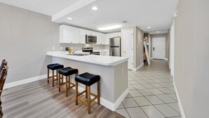 A modern kitchen with white cabinetry, a central island with bar stools, and a visible microwave. - Amalfi Coast - Spacious Condo! (Miramar Beach)