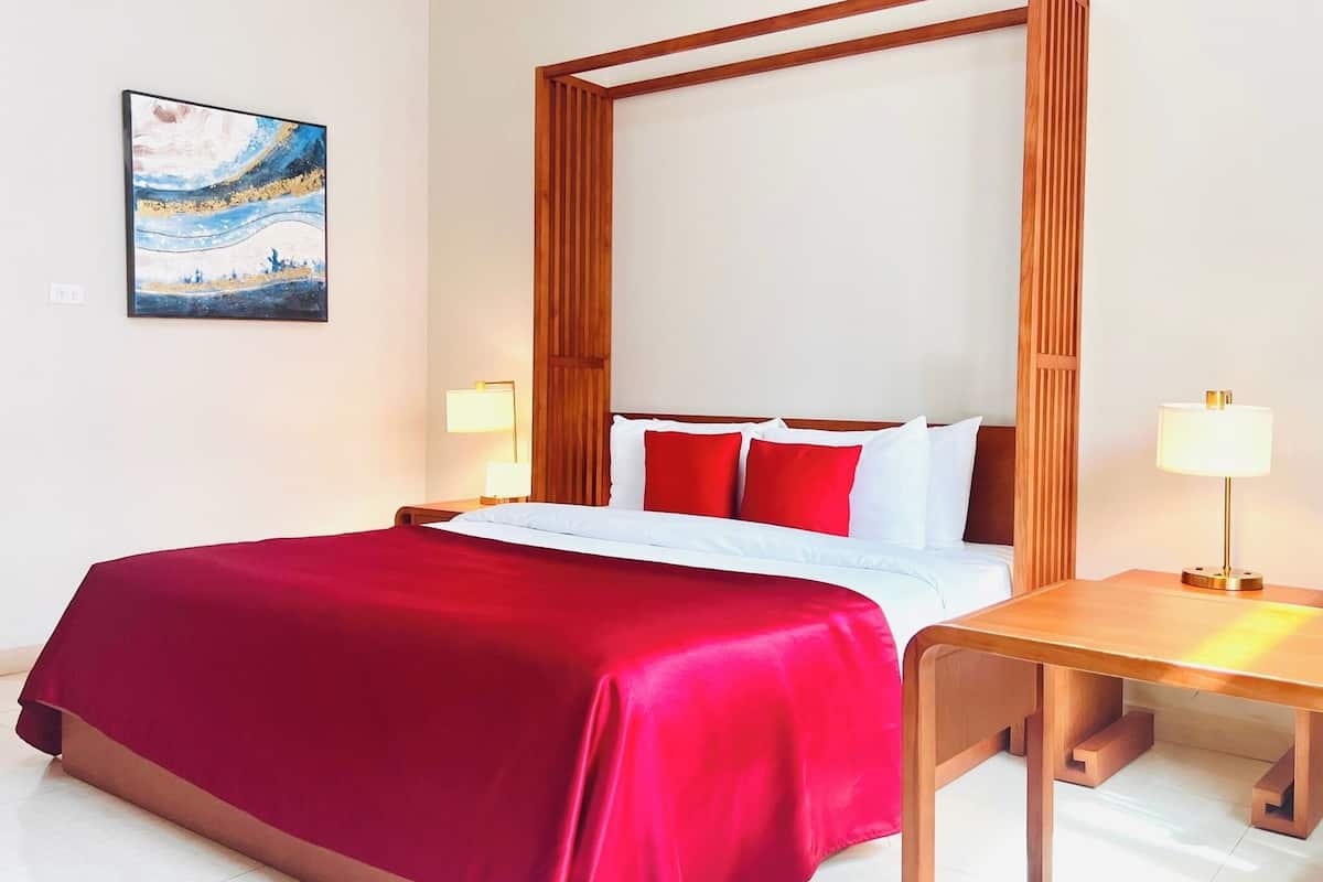 A hotel room with a large bed, red bedding, wooden headboard, bedside tables with lamps, and a painting on the wall.