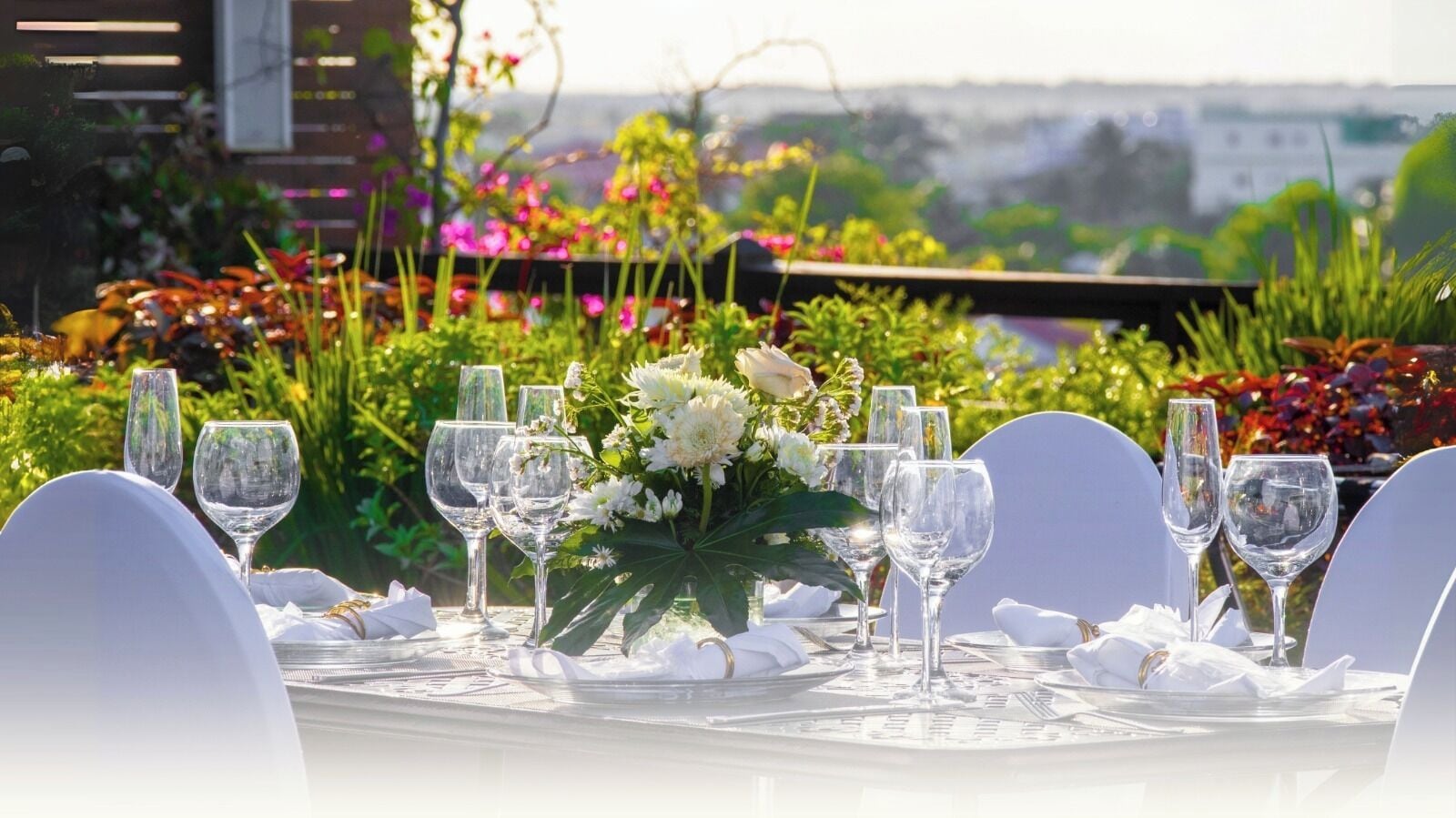A table set for a formal outdoor dining event with white chairs and clear glassware, surrounded by lush greenery and flowers.