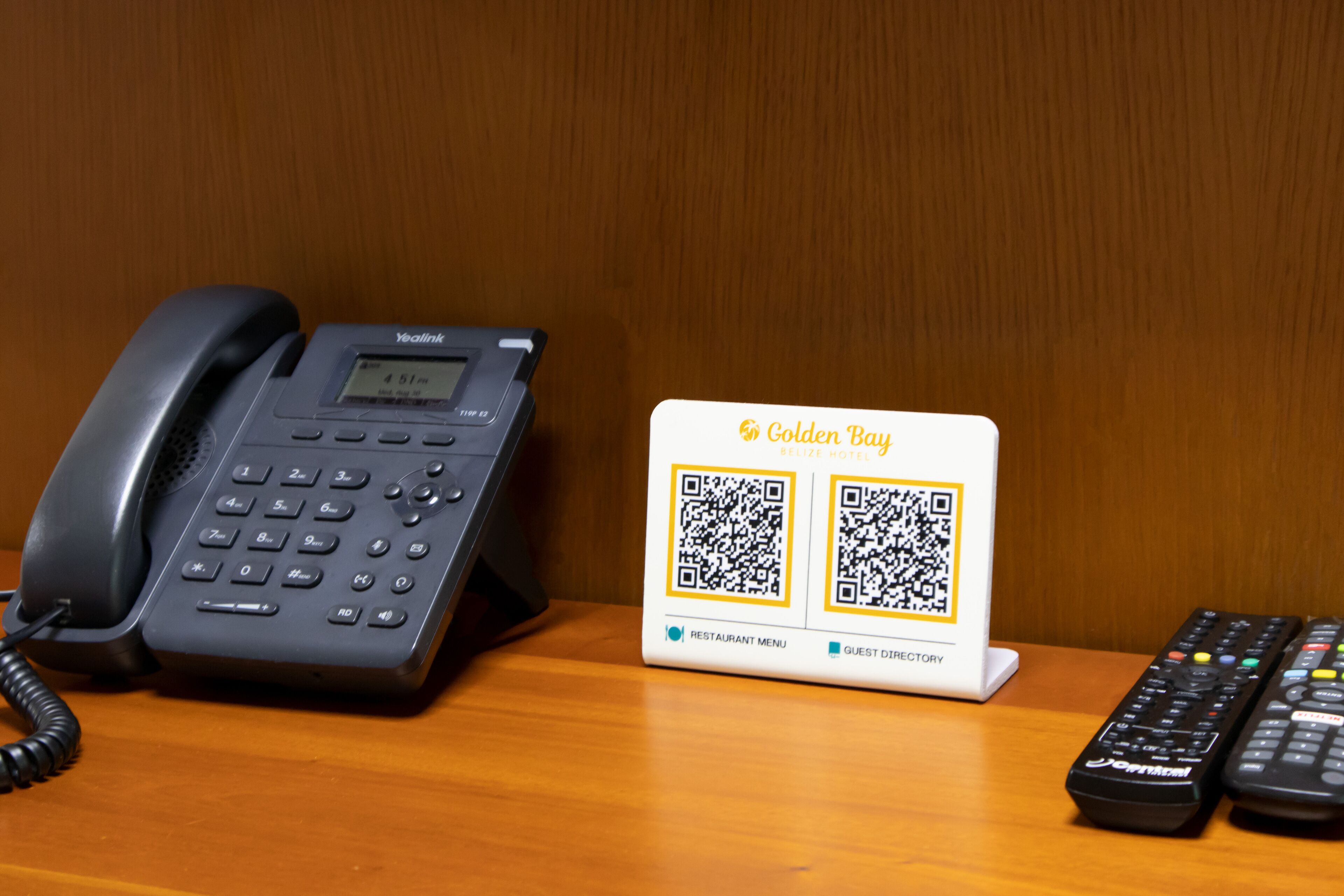 A hotel room with a desk, a telephone, a QR code stand, and two remote controls.