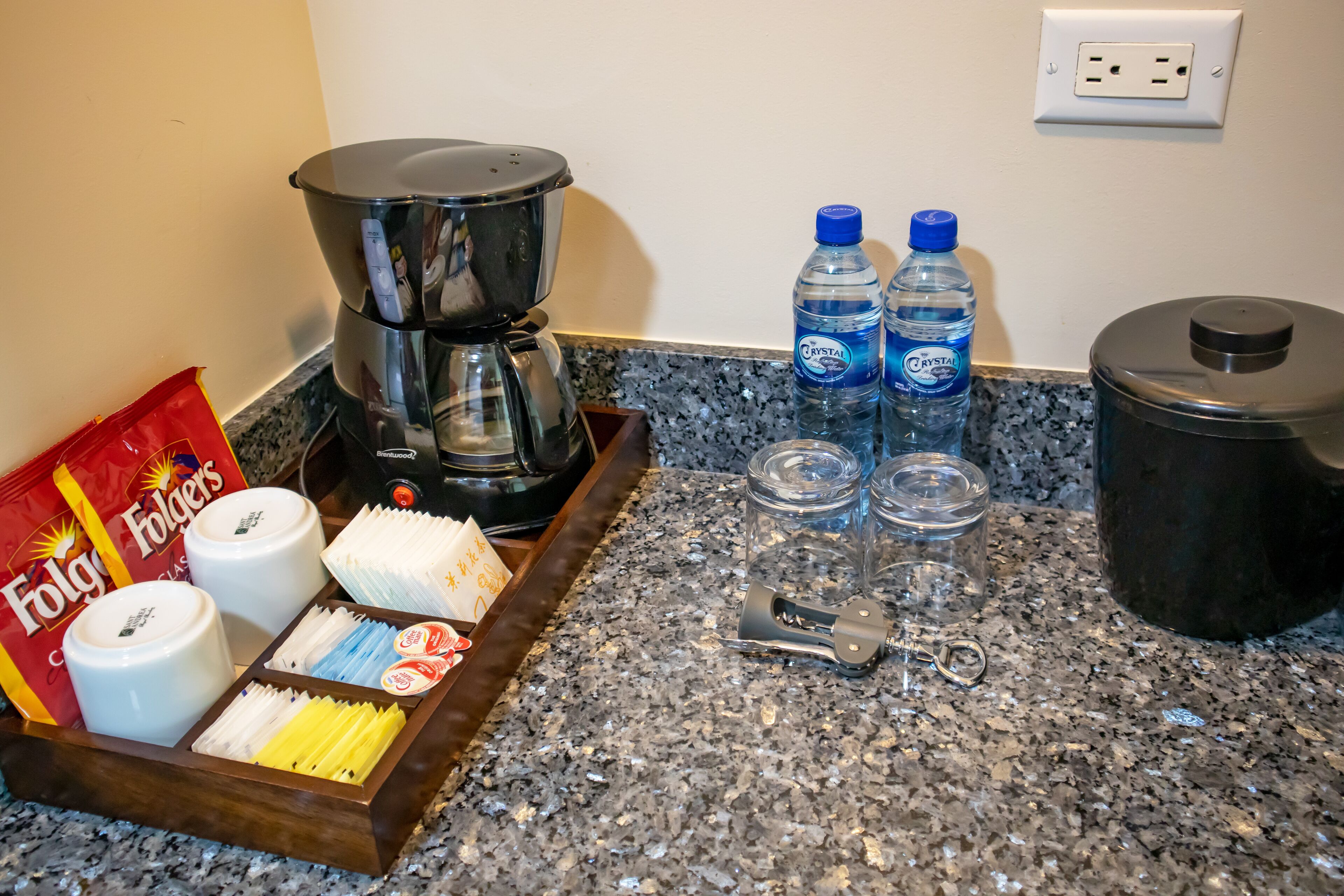 Una encimera de cocina con una cafetera, dos botellas de agua, un tarro, un cuchillo y un paquete de café Folgers.