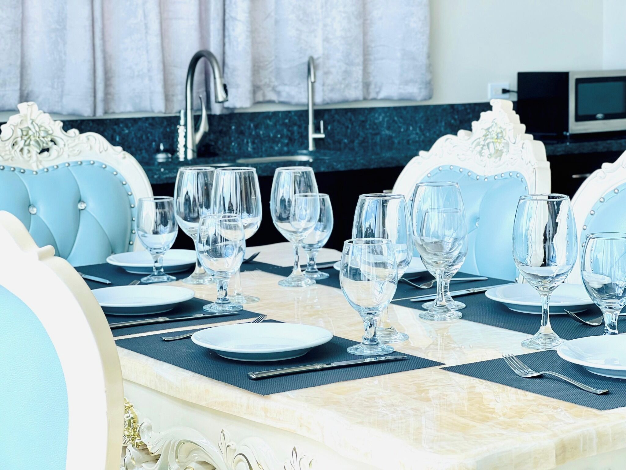 Une table à manger dressée avec de nombreux verres à vin, des assiettes et des couverts, entourée de chaises rembourrées ornées d’un motif capitonné.