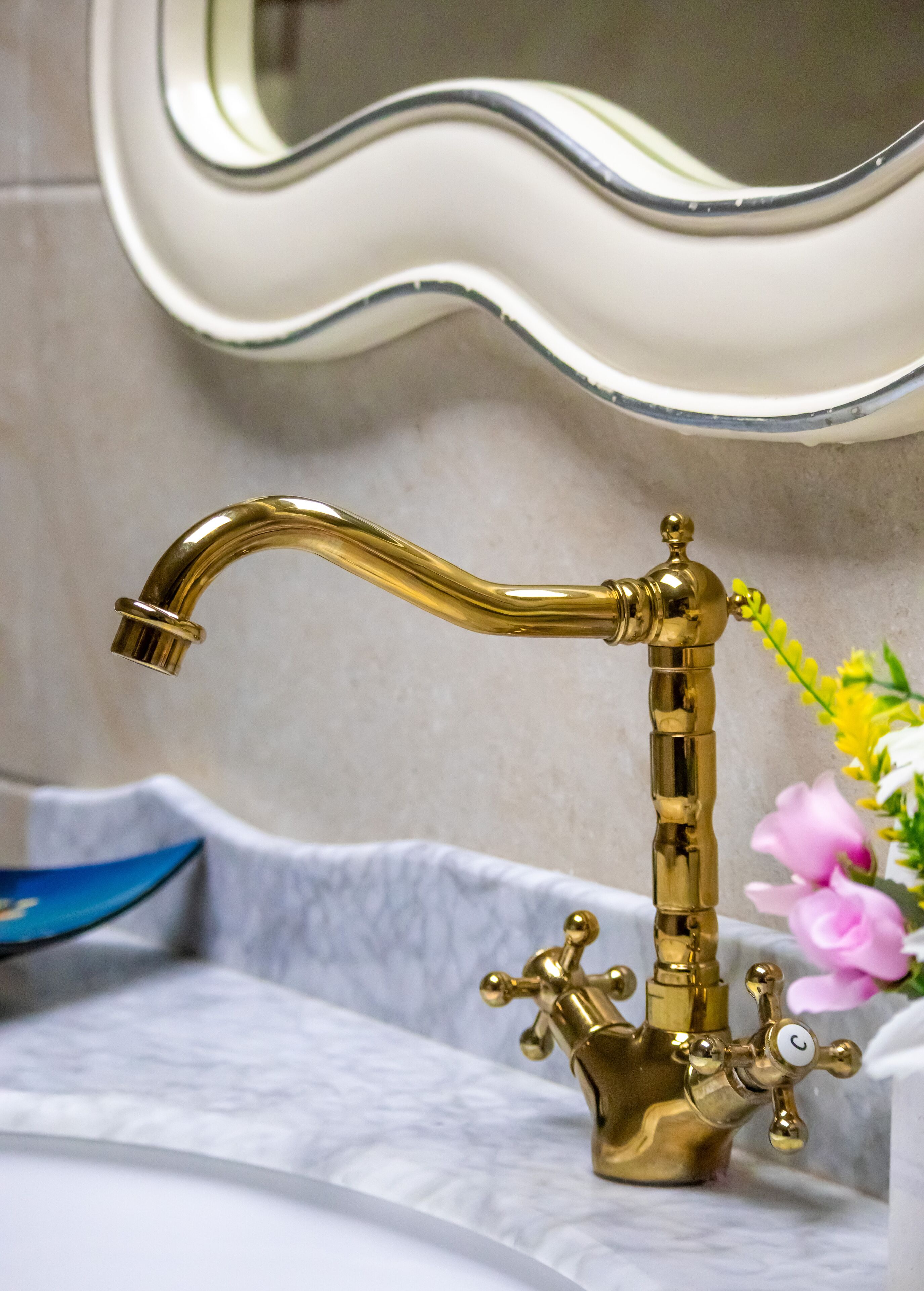 Un baño con grifo dorado, lavamanos blanco y espejo de borde ondulado.