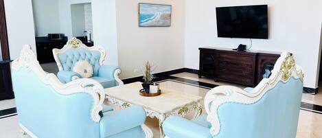 A spacious living room with a light blue sofa set, a marble coffee table, and a dark wooden cabinet with a flat-screen TV mounted above it.