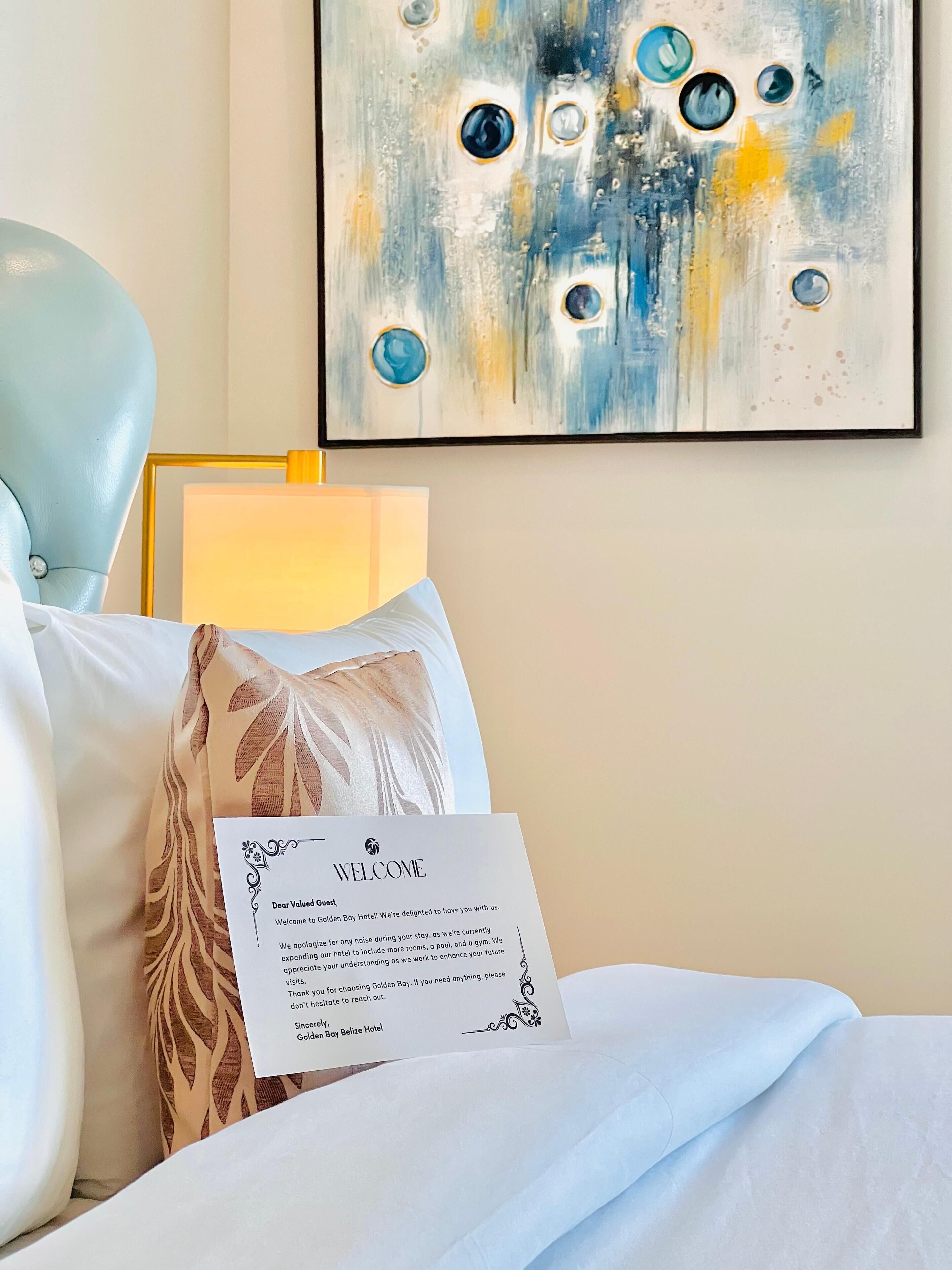 Een hotelkamer met een bed, een kussen met bladmotief, een welkomstkaart, een lamp en een kunstwerk aan de muur.