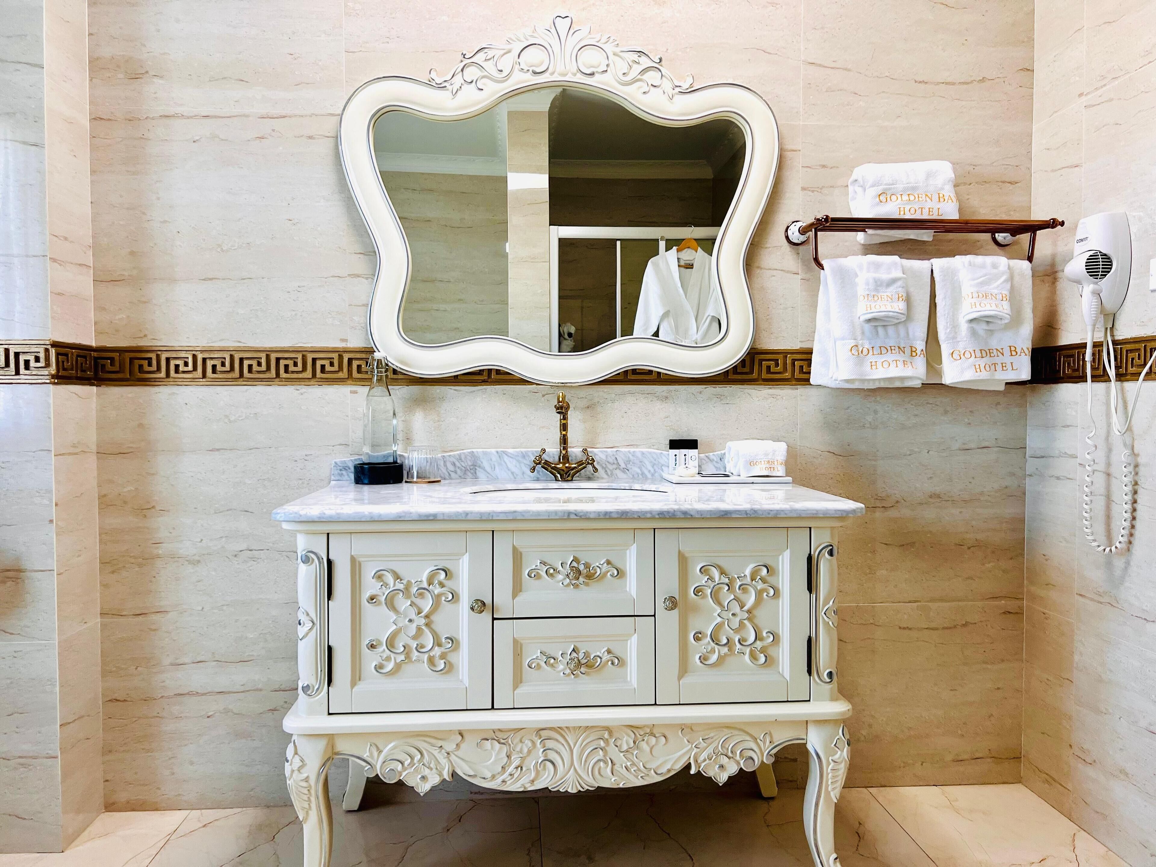 A bathroom with a marble sink, ornate cabinet, and a large mirror. There are towels hanging on a rack and a hairdryer on the wall.