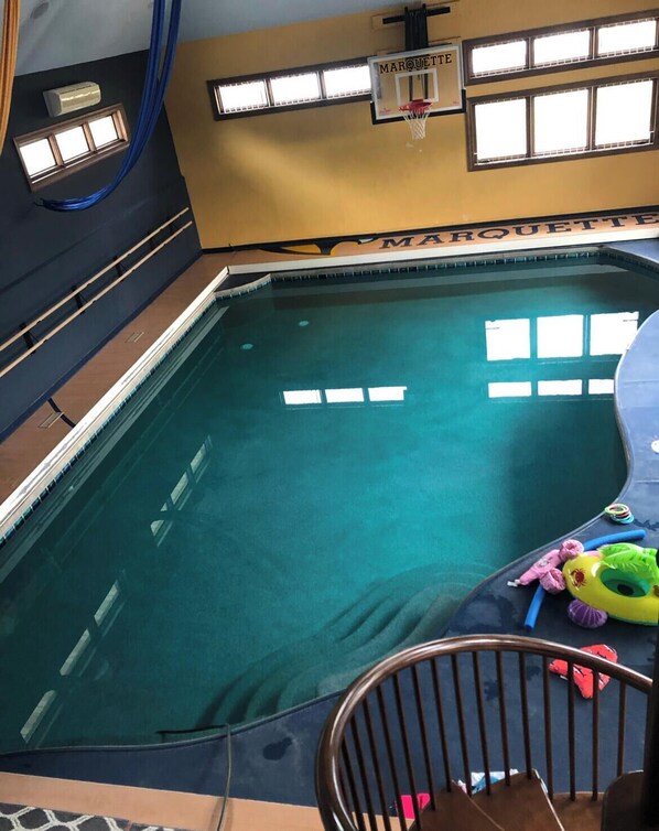 An indoor swimming pool with clear water, a basketball hoop, and a window with a view.