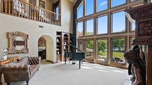 A spacious living room with a grand piano, a brown leather sofa, and a large window offering a view of the outdoors. - Amazing Luxury Mansion ~ Indoor Pool ~ Stunning Lake Views ~ One Of A Kind! (Newton)
