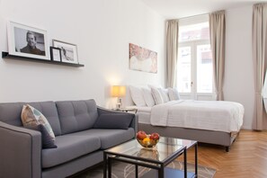 Una habitación de hotel moderna con un sofá gris, una mesa de centro de vidrio con fruta, una cama con ropa de cama blanca y obras de arte en las paredes.