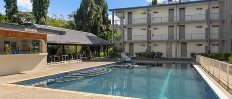 A swimming pool with a slide, surrounded by a multi-story building with balconies, and a covered area with seating.