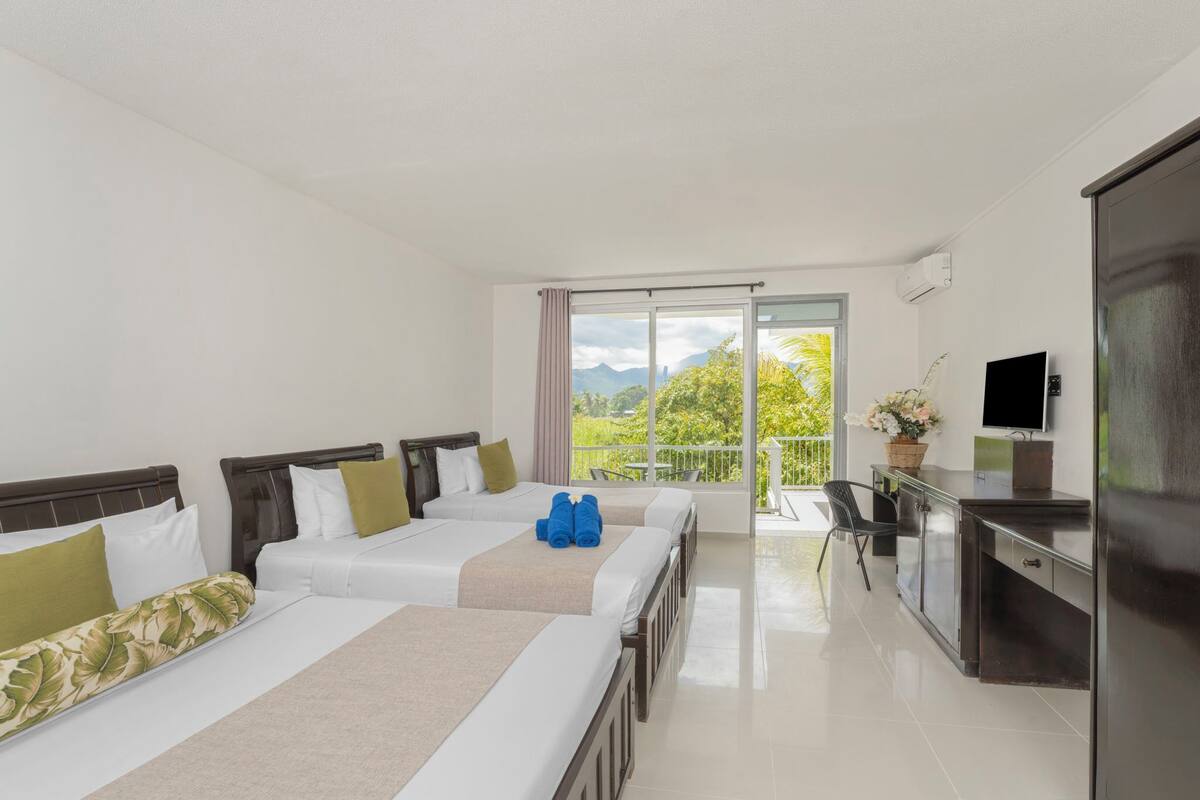 Quarto de hotel com duas camas, uma escrivaninha, televisão e varanda com vista para vegetação e montanhas.