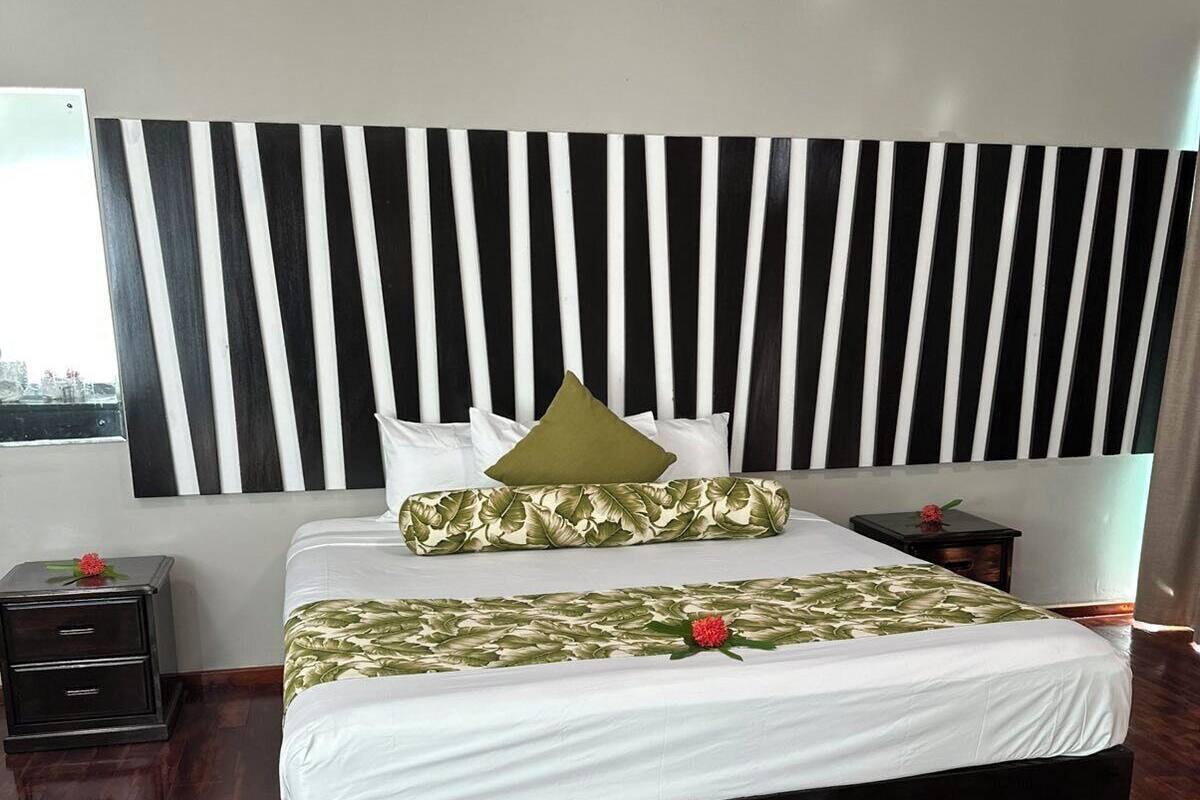 Quarto com cabeceira listrada em preto e branco, cama com roupa de cama branca e almofadas estampadas com folhas, mesa de cabeceira com uma flor e vista da cidade pela janela.