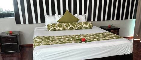 A bedroom with a black and white striped headboard, a bed with white bedding and green leaf-patterned pillows, a nightstand with a flower, and a view of the city through a window.