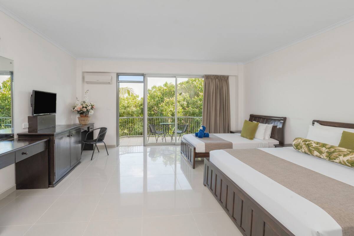 Quarto de hotel com cama, escrivaninha, televisão e varanda com vista para vegetação.