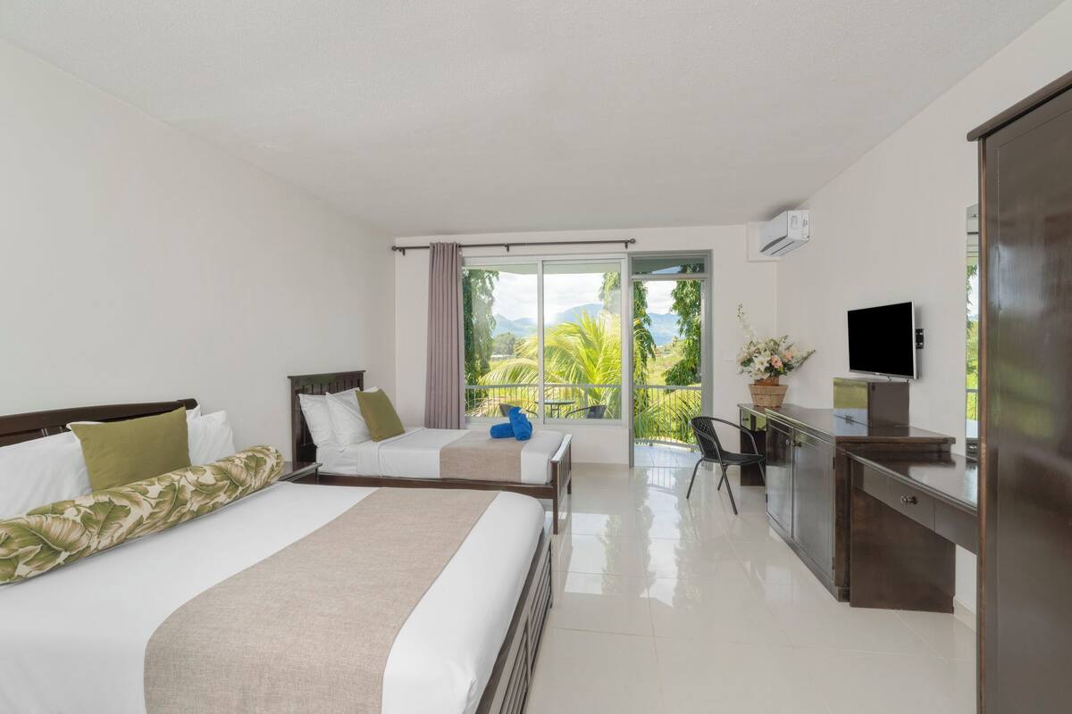 Quarto de hotel com duas camas, uma escrivaninha, televisão e varanda com vista para o verde.