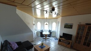 A living room with a black sofa, a wooden coffee table, a TV stand, and a dining area with a table and chairs. - Vacation apartment type 1, 50sqm, 1 bedroom - Ferienhaus Irene (Vogtsburg im Kaiserstuhl)
