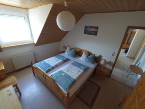 Ein Schlafzimmer mit schrägen Wänden, einem großen Bett mit blauer Bettwäsche, einem hölzernen Kopfteil, einem Nachttisch mit Lampe und einem Fenster mit Jalousien.