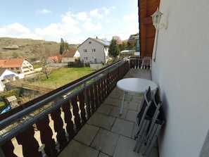 Un balcon avec une petite table et des chaises, d’où l’on peut admirer des maisons et de la verdure en arrière-plan.