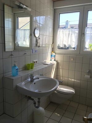 Une salle de bain avec un lavabo blanc, un miroir et des toilettes. Il y a une fenêtre donnant sur les bâtiments à l’extérieur.