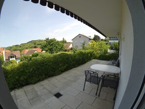 Eine Terrasse mit einem weißen Tisch und zwei Stühlen, einem Metallgrill und Blick auf eine Hügellandschaft mit Häusern.
