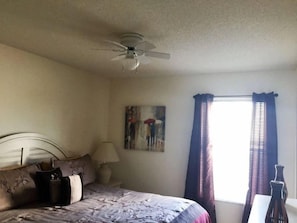A bedroom with a bed, a ceiling fan, a painting, a window with curtains, and a vase on a nightstand.
