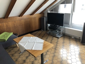 A room with a sloped ceiling, wooden panel walls, a tiled floor, a wooden table with a white cloth, a gray sofa, a television on a stand, and a radiator.