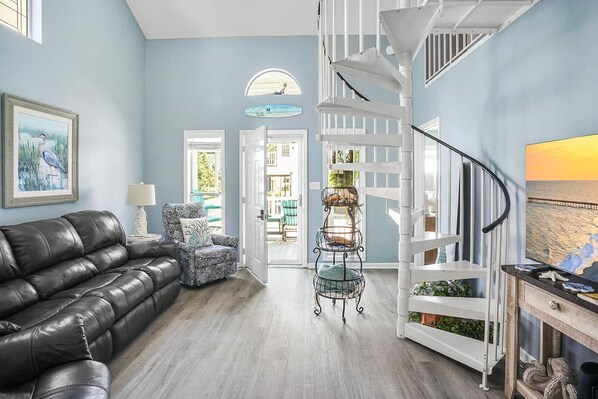 A spacious living room with a black leather sofa, a patterned armchair, a metal birdcage, and a white staircase.
