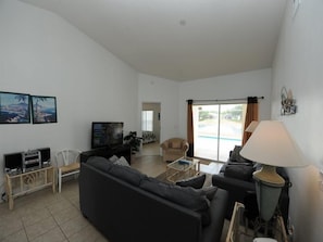 A living room with a sofa, TV, and a lamp. There is a window with curtains and a view of a pool.