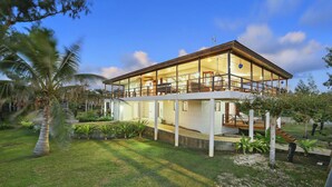 Une maison moderne à deux étages, dotée d’un grand balcon et entourée d’un jardin bien entretenu, agrémenté de palmiers et d’autres plantes.