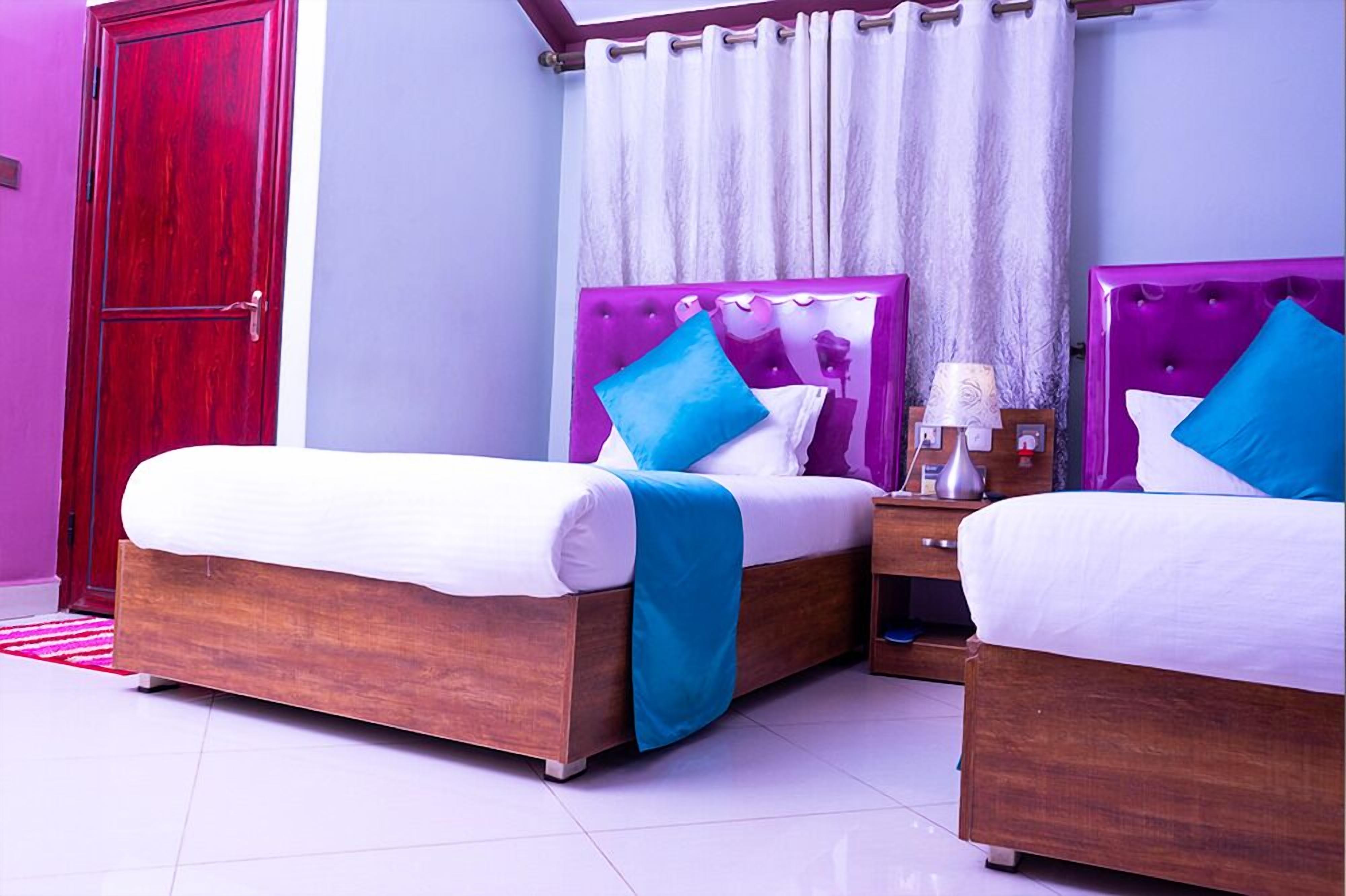 A hotel room with two beds, a wooden wardrobe, a nightstand with a lamp, and a window with curtains.
