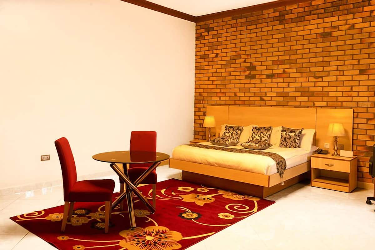 A bedroom with a brick wall, a bed with patterned pillows, a red chair, a small round table, and a nightstand with a lamp.