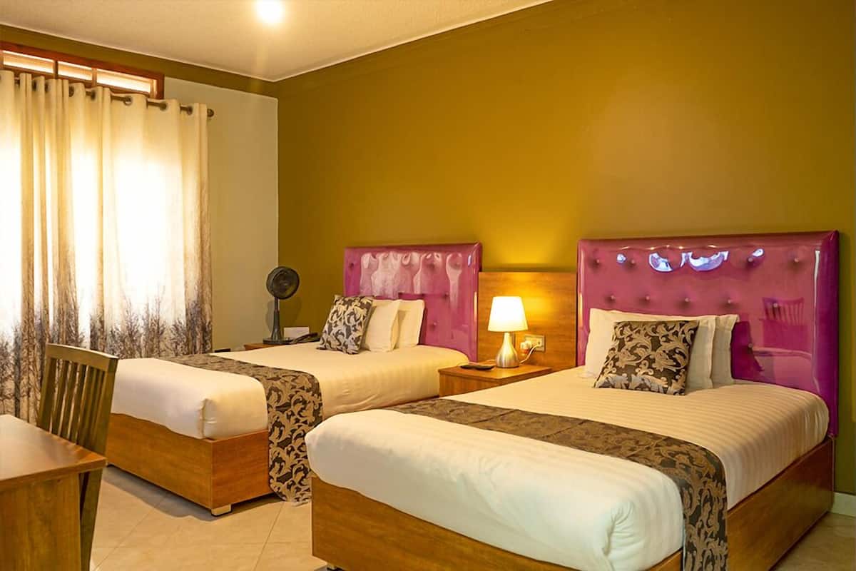 A hotel room with two beds, a wooden chair, a bedside table with a lamp, and a window with curtains.