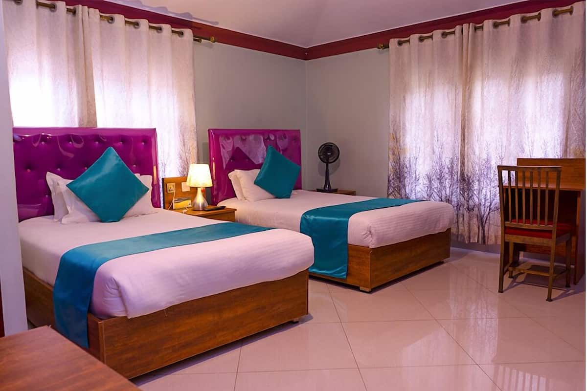 A hotel room with two beds, each with a wooden frame and teal bedspreads. There is a wooden chair and a bedside table with a lamp.