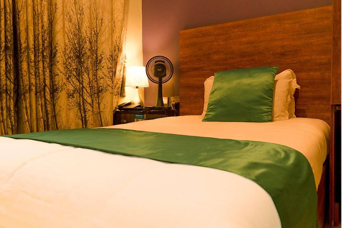 A neatly made bed with a green bedspread, a wooden headboard, a nightstand with a lamp and a fan, and a curtain with a tree pattern.