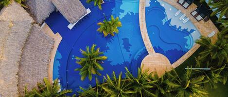 Vista aérea de una piscina con varias secciones, rodeada de palmeras y sillones de descanso.