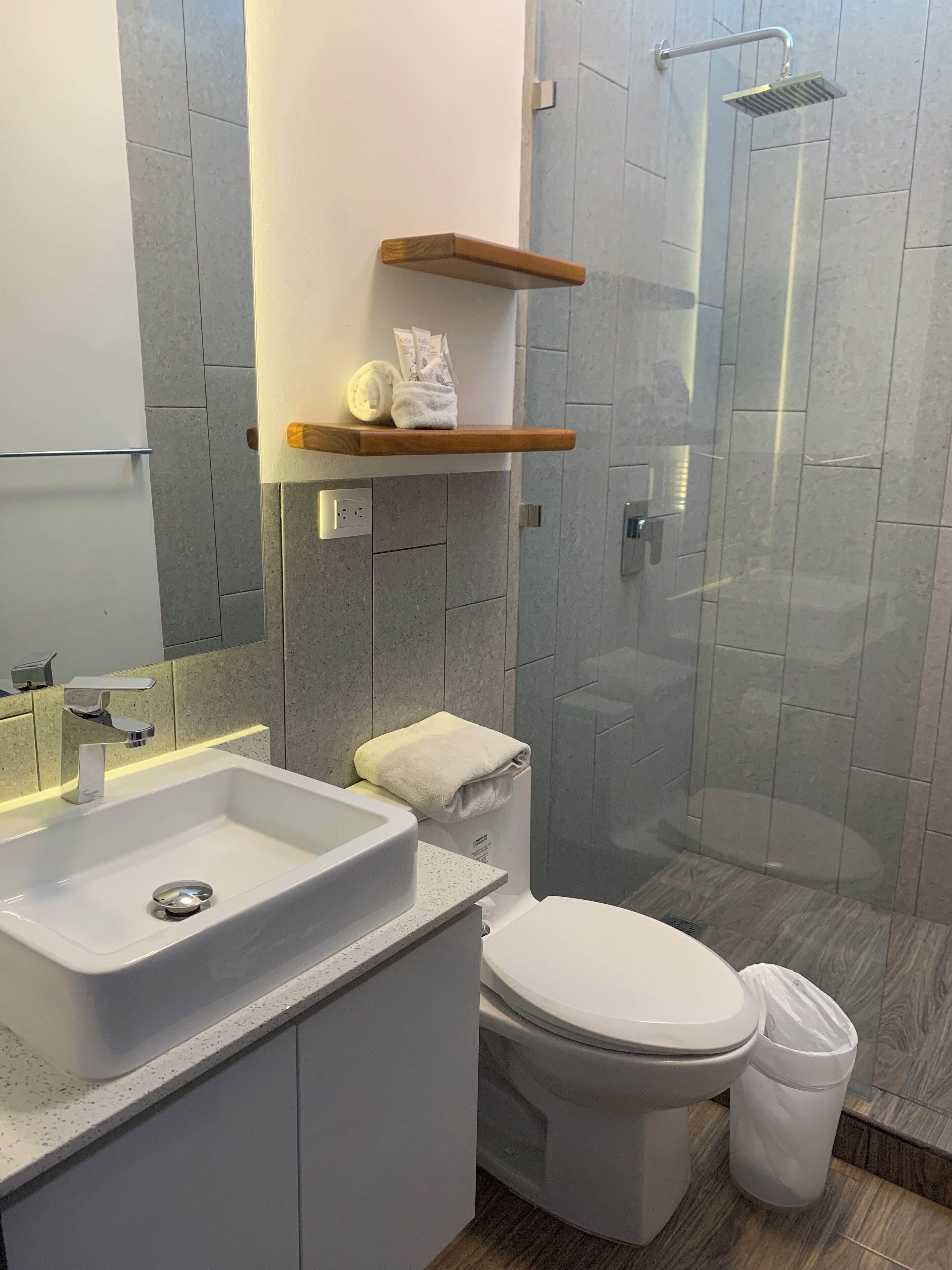 A modern bathroom with a white sink, toilet, and shower. There are wooden shelves above the toilet and a towel hanging on the wall.
