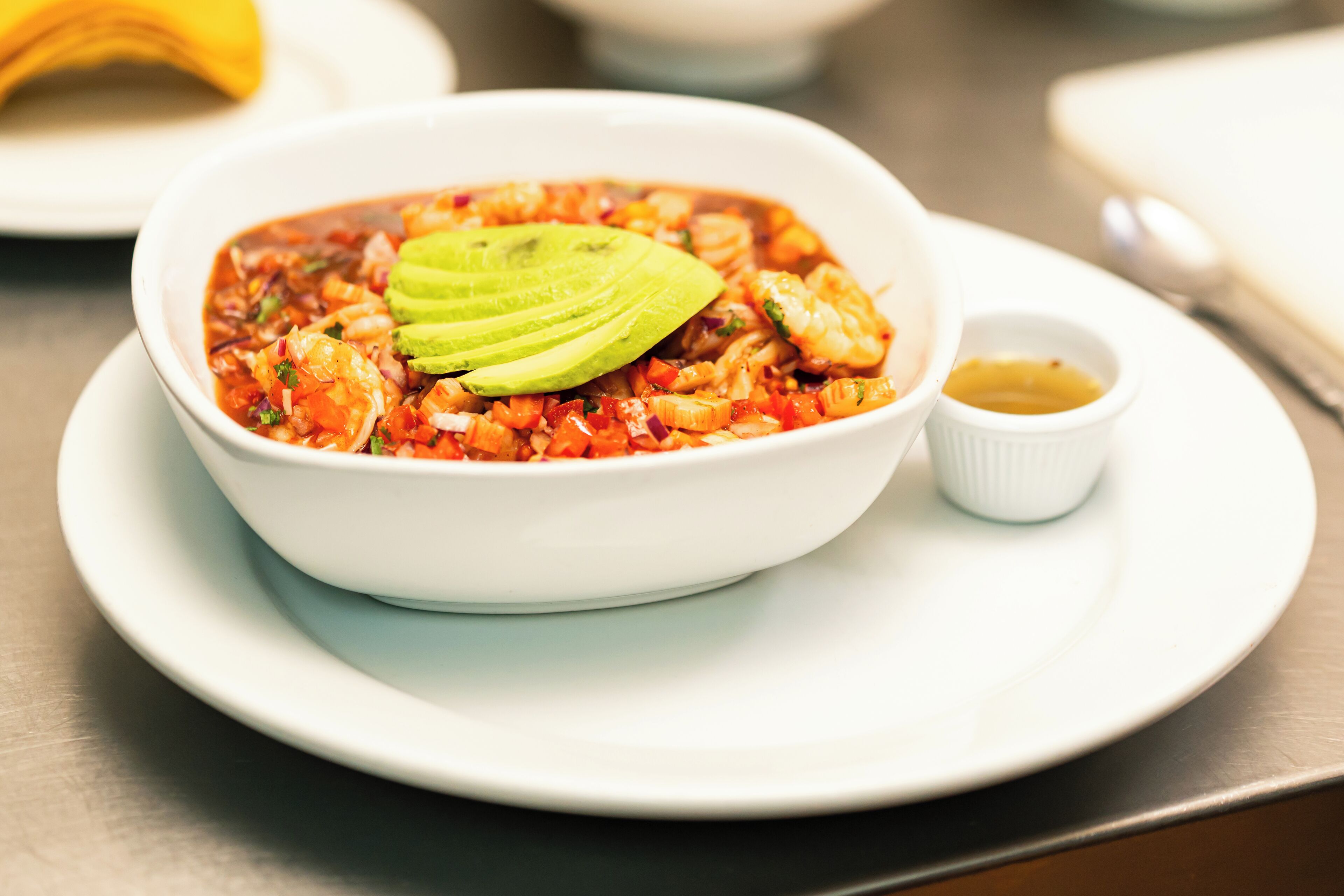 A bowl of soup with avocado slices on top, served on a white plate with a small bowl of sauce beside it.