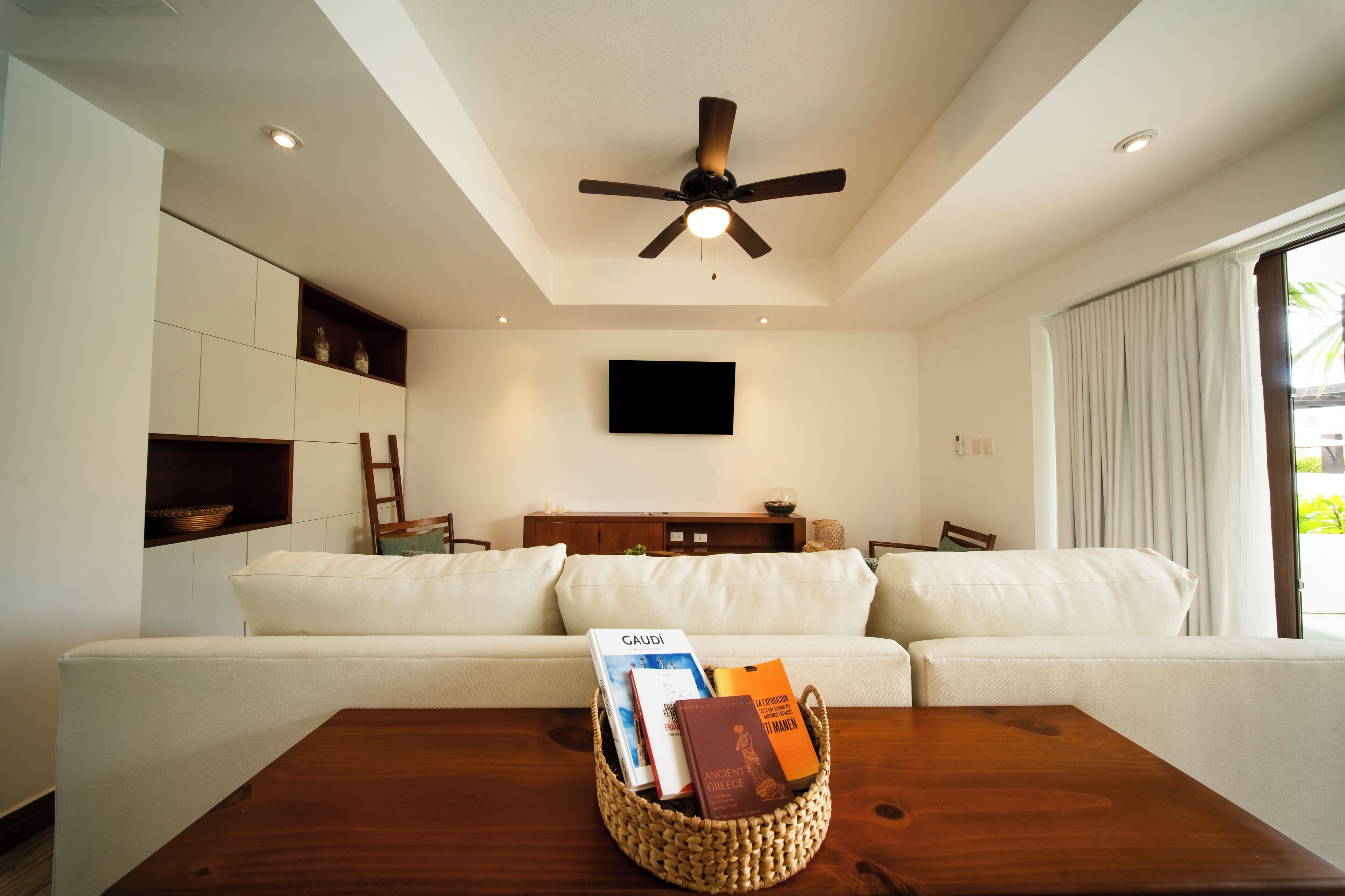 A modern living room with a white sofa, a wooden coffee table with books, a ceiling fan, and a flat-screen TV on the wall.