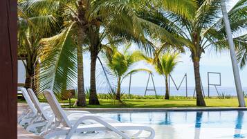 A poolside area with white lounge chairs, palm trees, and a view of the ocean.