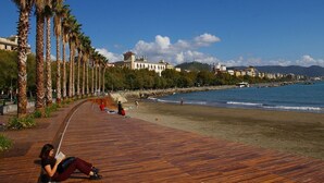 Une promenade en bois longeant une plage bordée de palmiers, avec un bâtiment en arrière-plan et un ciel bleu dégagé.