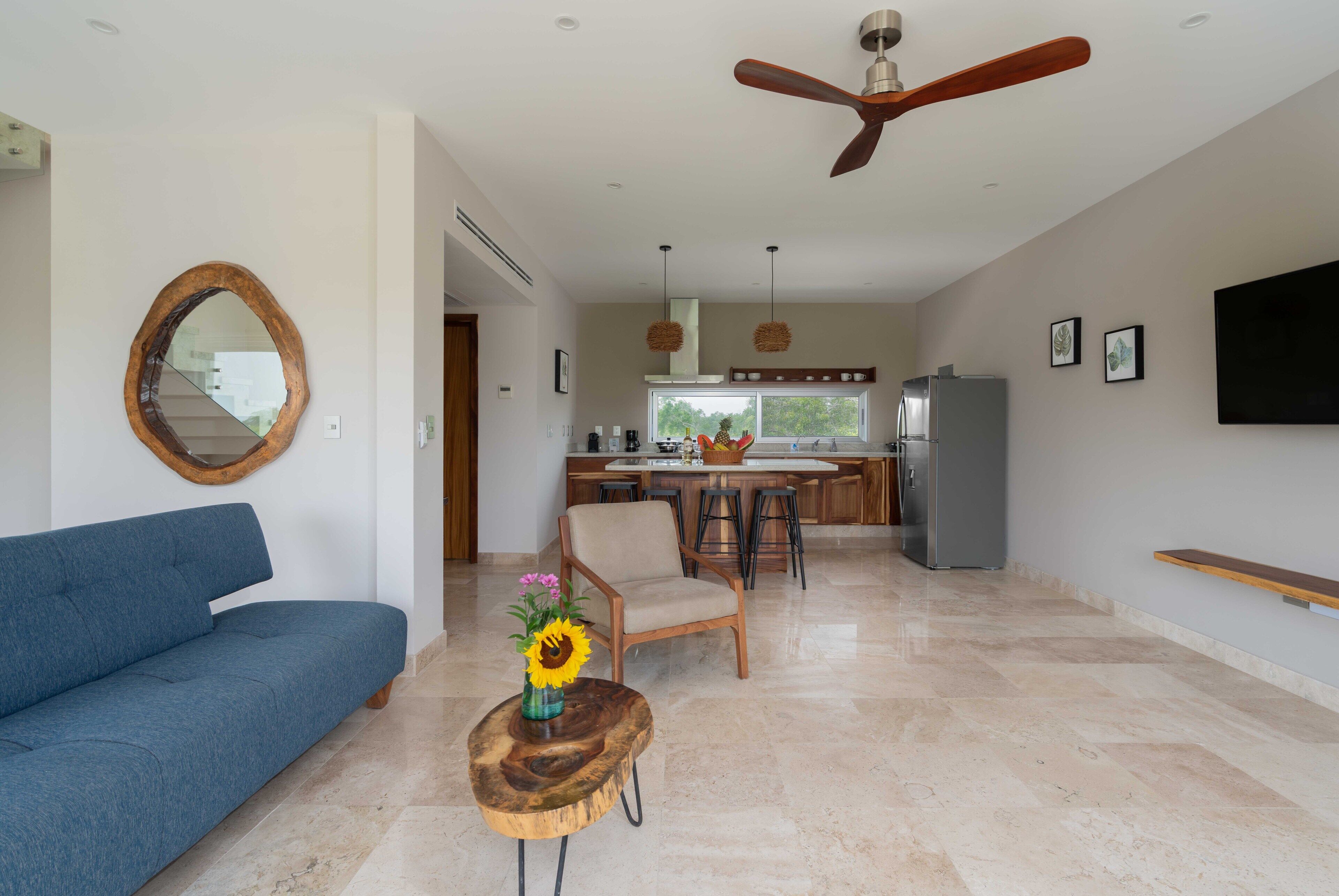 A spacious living room with a blue sofa, a wooden coffee table with a vase of flowers, a wooden chair, and a ceiling fan.