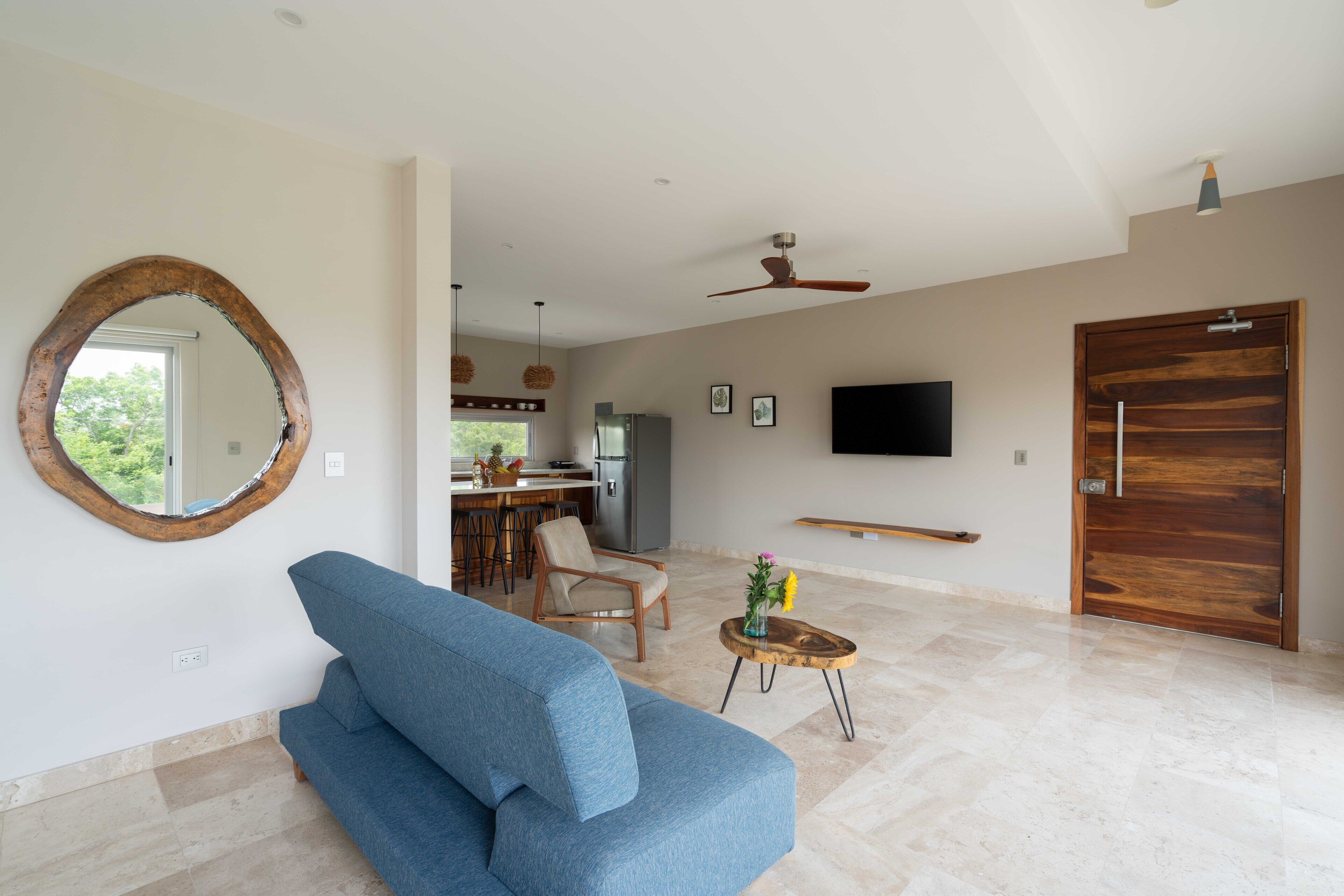 A modern living room with a blue sofa, a wooden-framed mirror, a ceiling fan, a TV, and a wooden door.