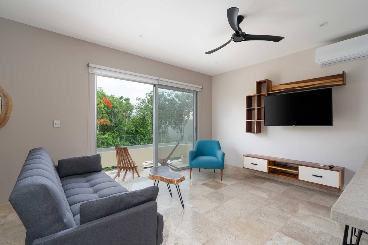 A modern living room with a grey sofa, a blue armchair, a wooden coffee table, a flat-screen TV mounted on the wall, and a ceiling fan.