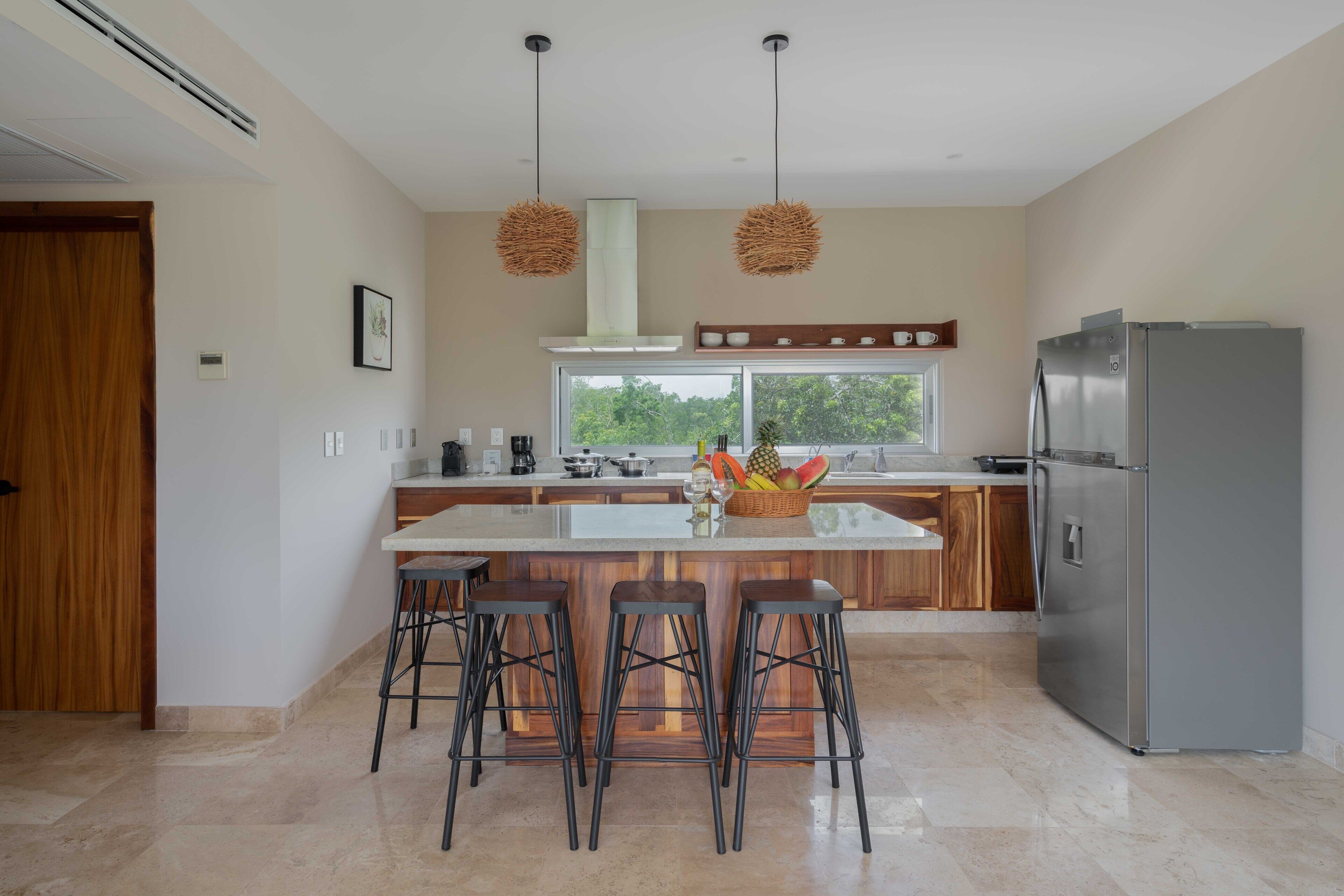 A modern kitchen with a central island, bar stools, a stainless steel refrigerator, and a large window with a view of greenery.
