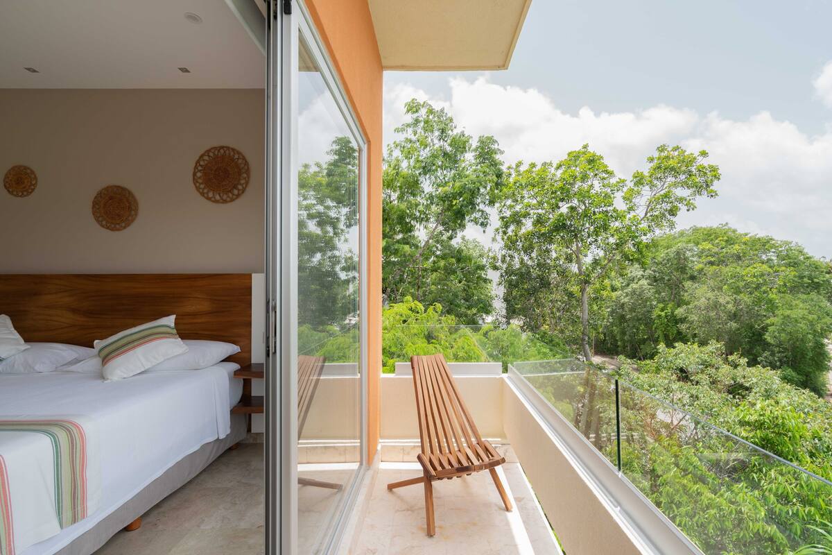 A bedroom with a large bed, a wooden chair, and a balcony with a view of trees.