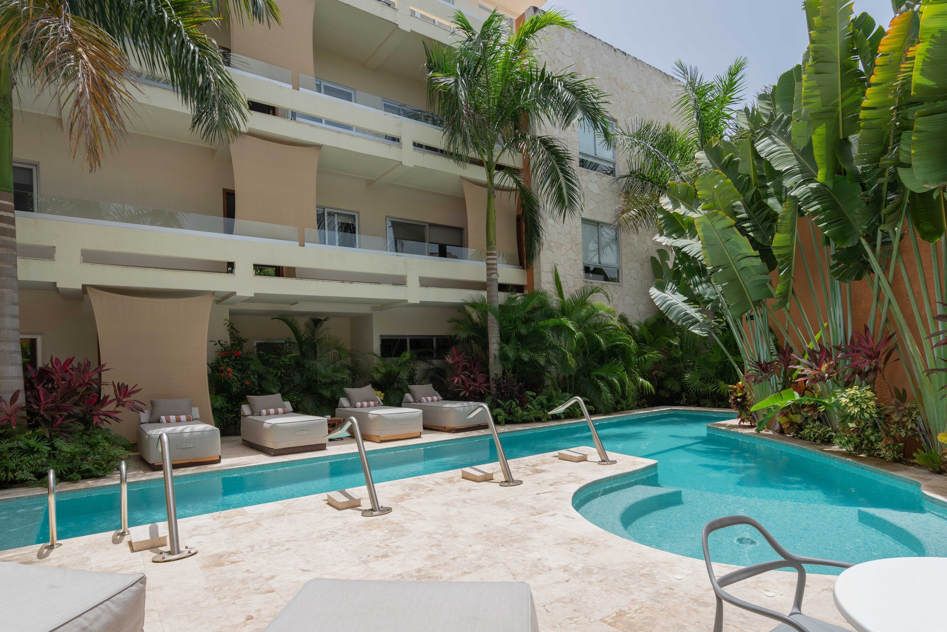 A pool area with lounge chairs and a small table, surrounded by palm trees and tropical plants.