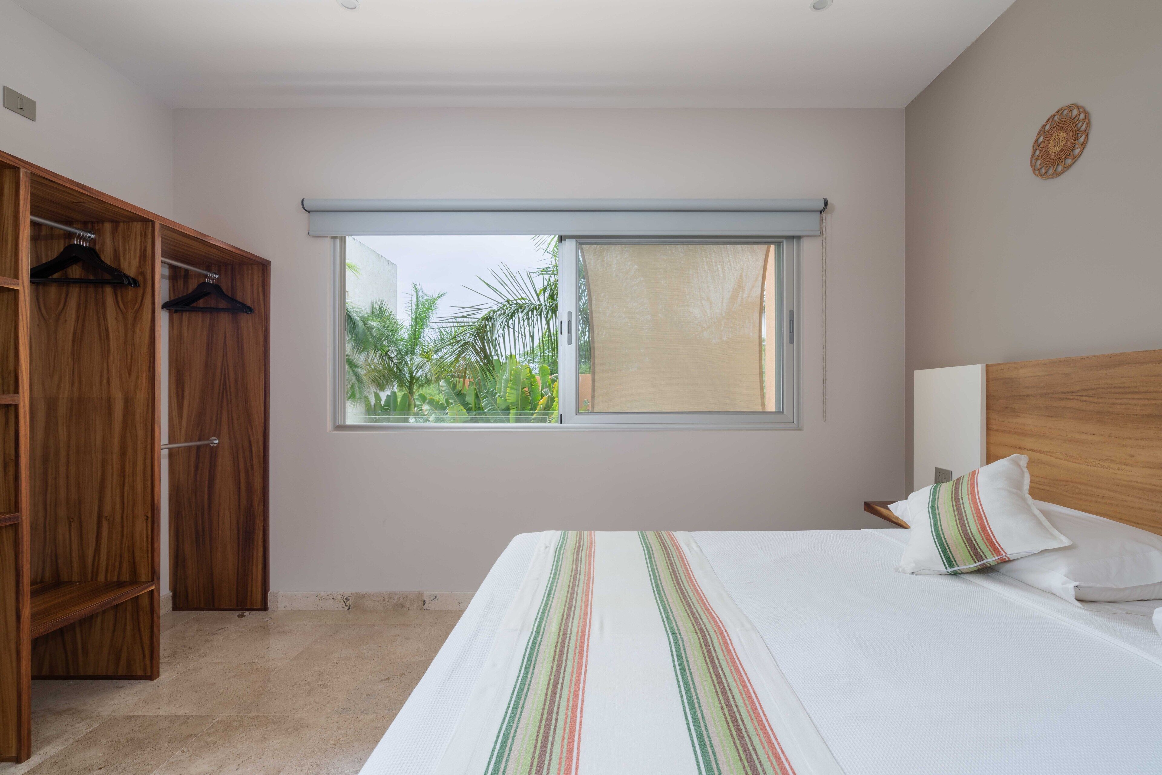 A bedroom with a bed, a wooden wardrobe, a window with a view of greenery, and a wall-mounted light fixture.