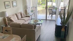Ein Wohnzimmer mit einer Couch, einem Glastisch und einem Esstisch mit Stühlen. Es gibt einen Balkon mit Blick auf Palmen und einen Pool.