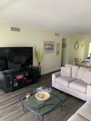 Una sala de estar con un televisor de pantalla plana, una mesa de centro de vidrio, un sofá color beige y una planta en maceta.